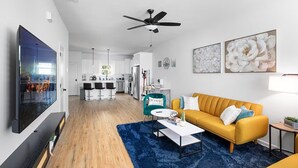 Een moderne woonkamer met een gele bank, een blauw vloerkleed en een keuken op de achtergrond.