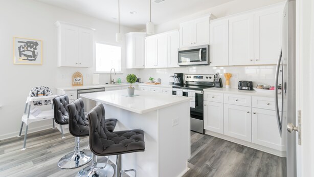 Een moderne keuken met witte kasten, een centraal eiland met barstoelen, roestvrijstalen apparatuur en een eethoek met een kinderstoel.
