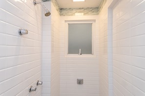 Une salle de bain avec des murs recouverts de carreaux blancs, une fenêtre et une douche équipée d’un pommeau de douche à main.