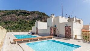 A rooftop swimming pool with clear blue water, surrounded by white buildings with red-tiled accents, set against a backdrop of a mountainous landscape. - Sunset Urca - Conforto e lazer (Rio de Janeiro)