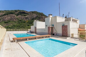 Une piscine sur le toit, avec une eau d’un bleu cristallin, entourée de bâtiments blancs rehaussés de tuiles rouges, le tout se détachant sur un paysage montagneux.