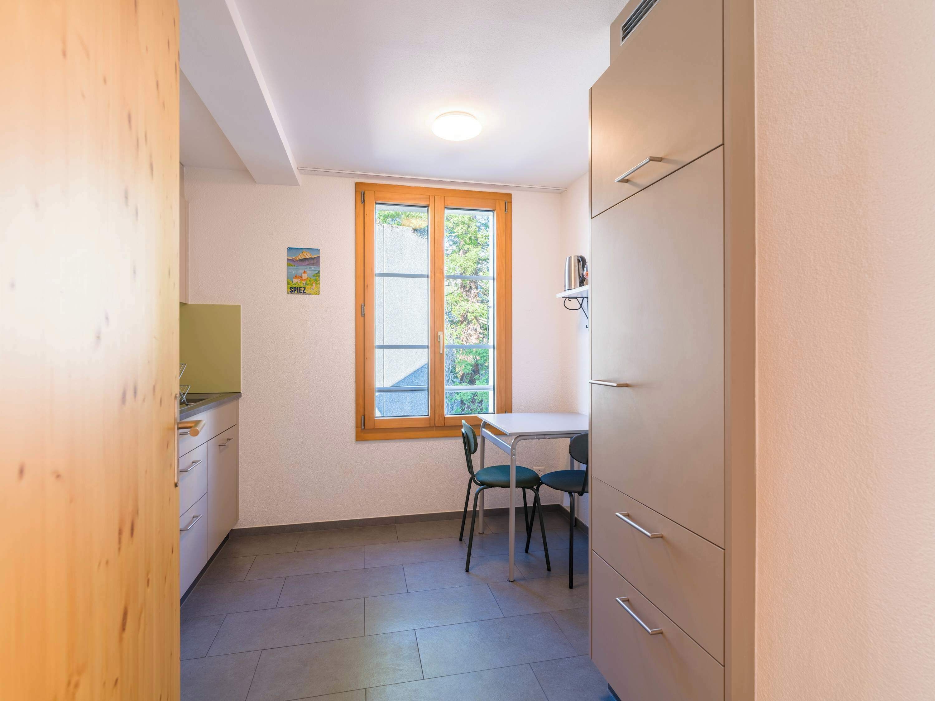 A small, bright room with a window, a table, two chairs, and a kitchenette.