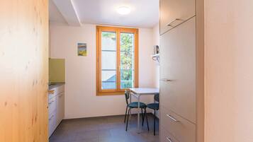 A small, bright room with a window, a table, two chairs, and a kitchenette.