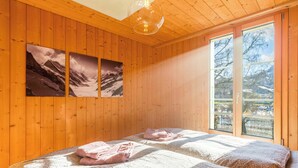 A wooden-paneled room with a bed, two wall-mounted pictures of mountains, and a large window with a view of trees and a building. - Lakefront Apartment in Spiez Bay (Spiez)