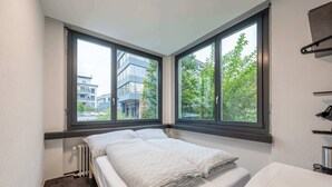 Een kleine, moderne slaapkamer met een bed, een radiator en een raam met uitzicht op gebouwen en bomen.
