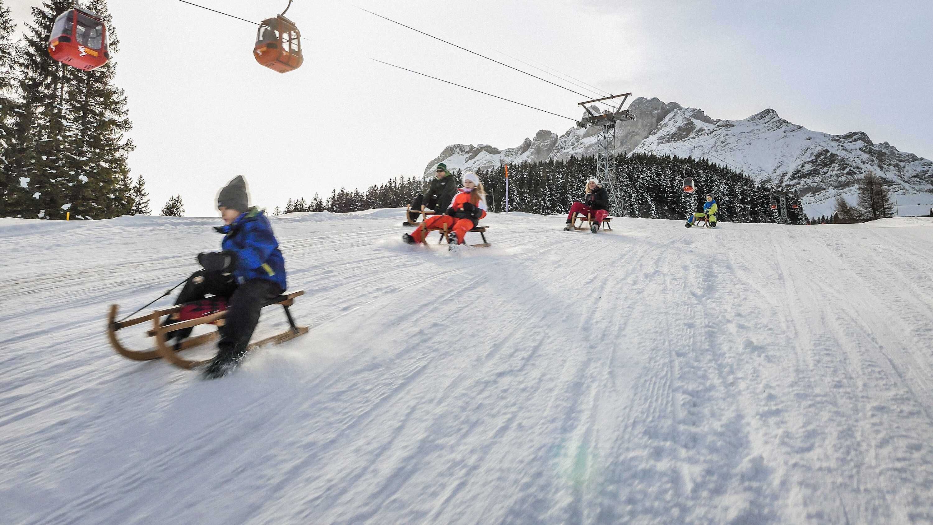 A snowy ski slope with people sledding and a cable car in the background.
