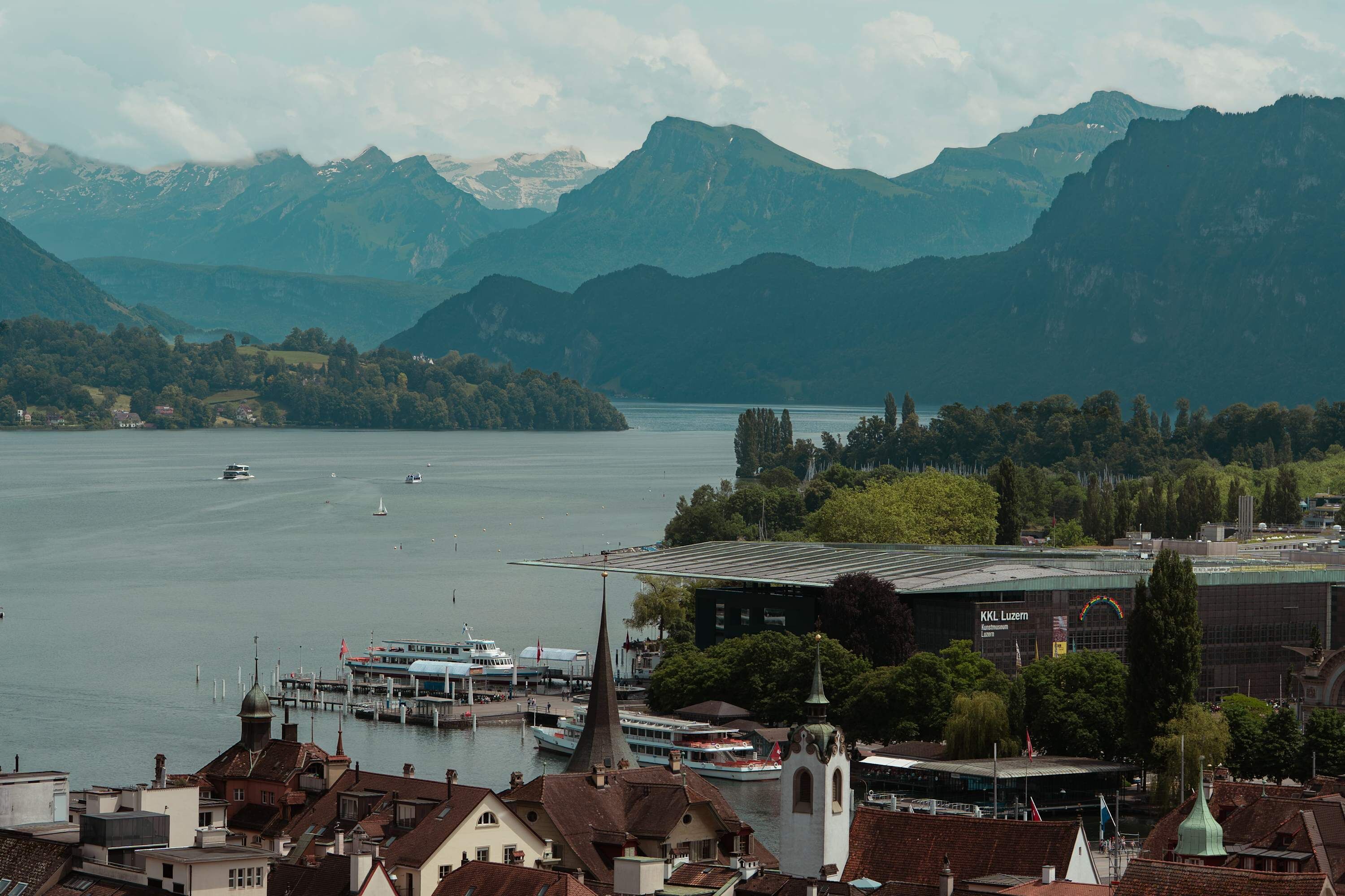 A lakeside town with a church, a dock with boats, and mountains in the background.