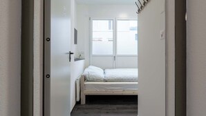 Passadís estret que condueix a una habitació amb un llit, una petita taula de nit i una finestra.