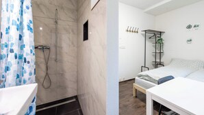 Een badkamer met een douche, toilet en wastafel. Er staat een bed met nachtkastje en stoel.