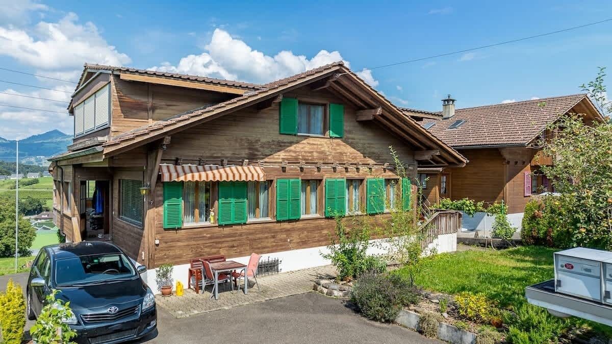 A wooden house with green shutters, a car parked in front, and a garden with plants and a small table.