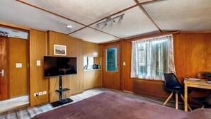 Una habitación compacta con paneles de madera, televisor, escritorio con silla y ventana con cortinas traslúcidas.