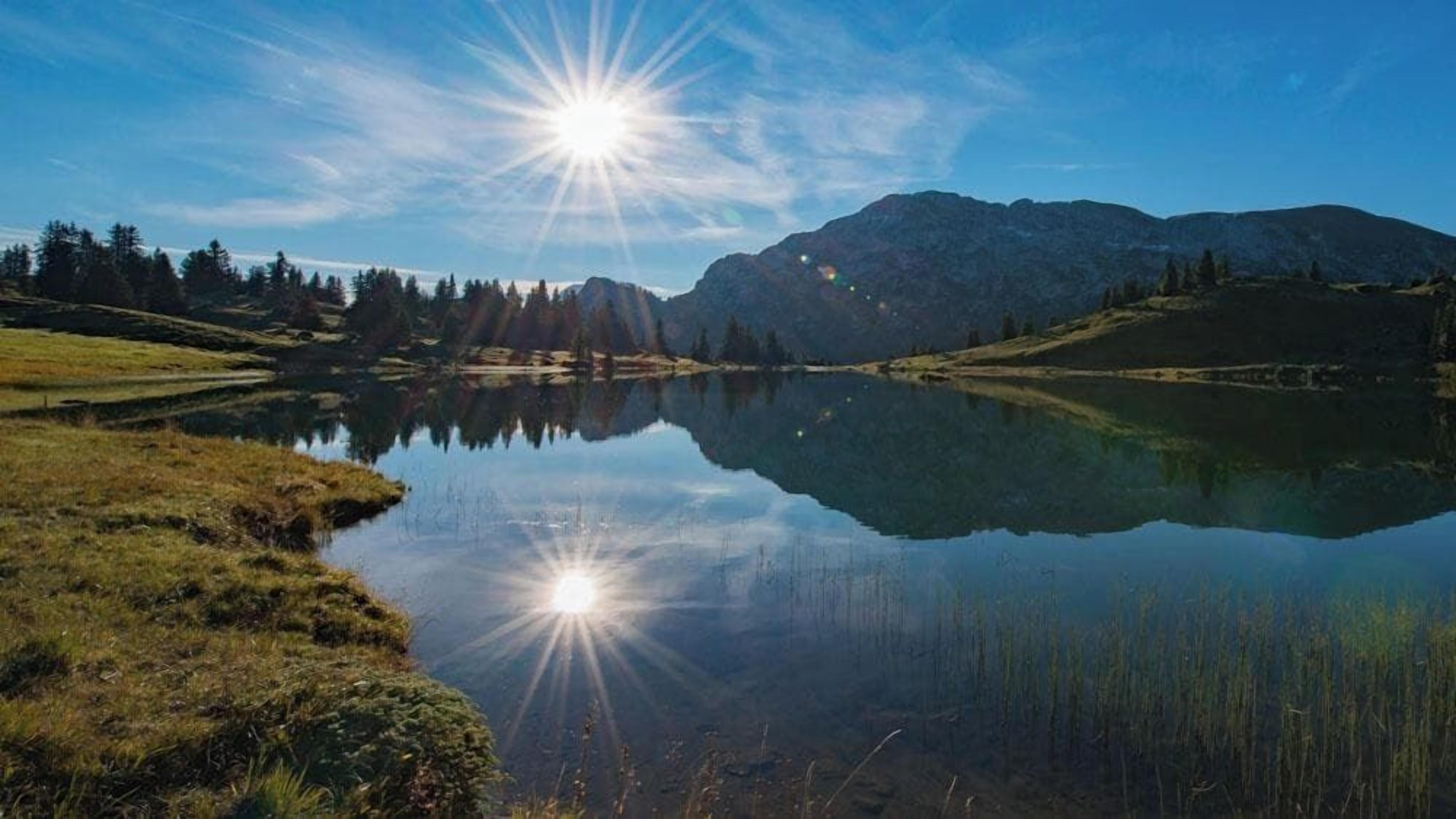 Ein ruhiger See mit Bergen im Hintergrund und hell scheinender Sonne.