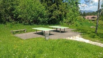 Un área de estar al aire libre con dos mesas verdes y un banco, rodeada de exuberante follaje y un cielo despejado.