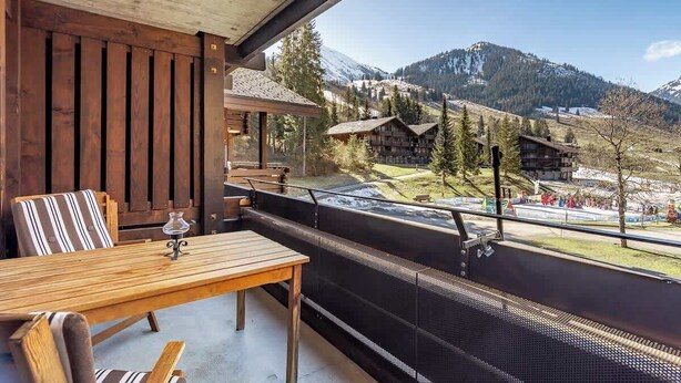 Un balcon avec une table et des chaises en bois, donnant sur un paysage montagneux parsemé de bâtiments et d’arbres.