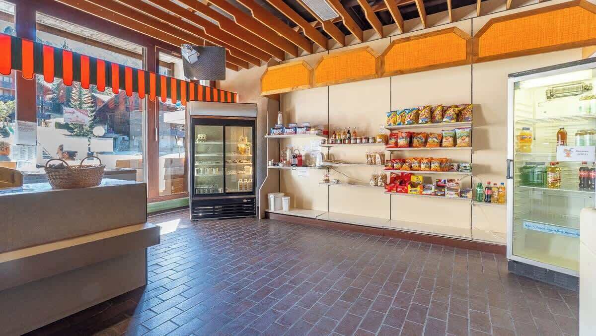Een restaurantinterieur met een stenen vloer, een toonbank met een mand, een koelkast vol drankjes en planken met snacks.