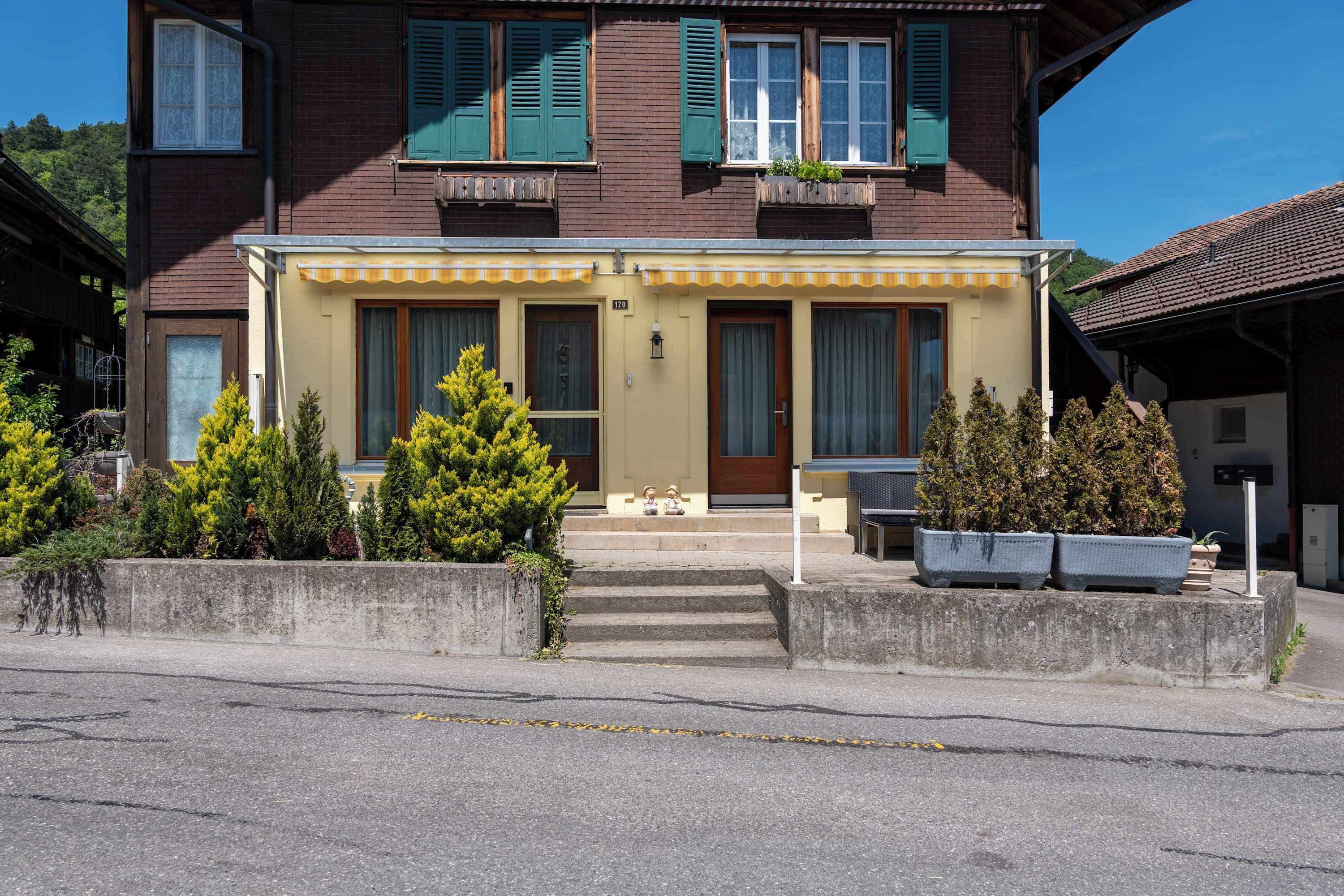 A two-story building with a yellow facade, green shutters, and a small porch with steps leading to the entrance.