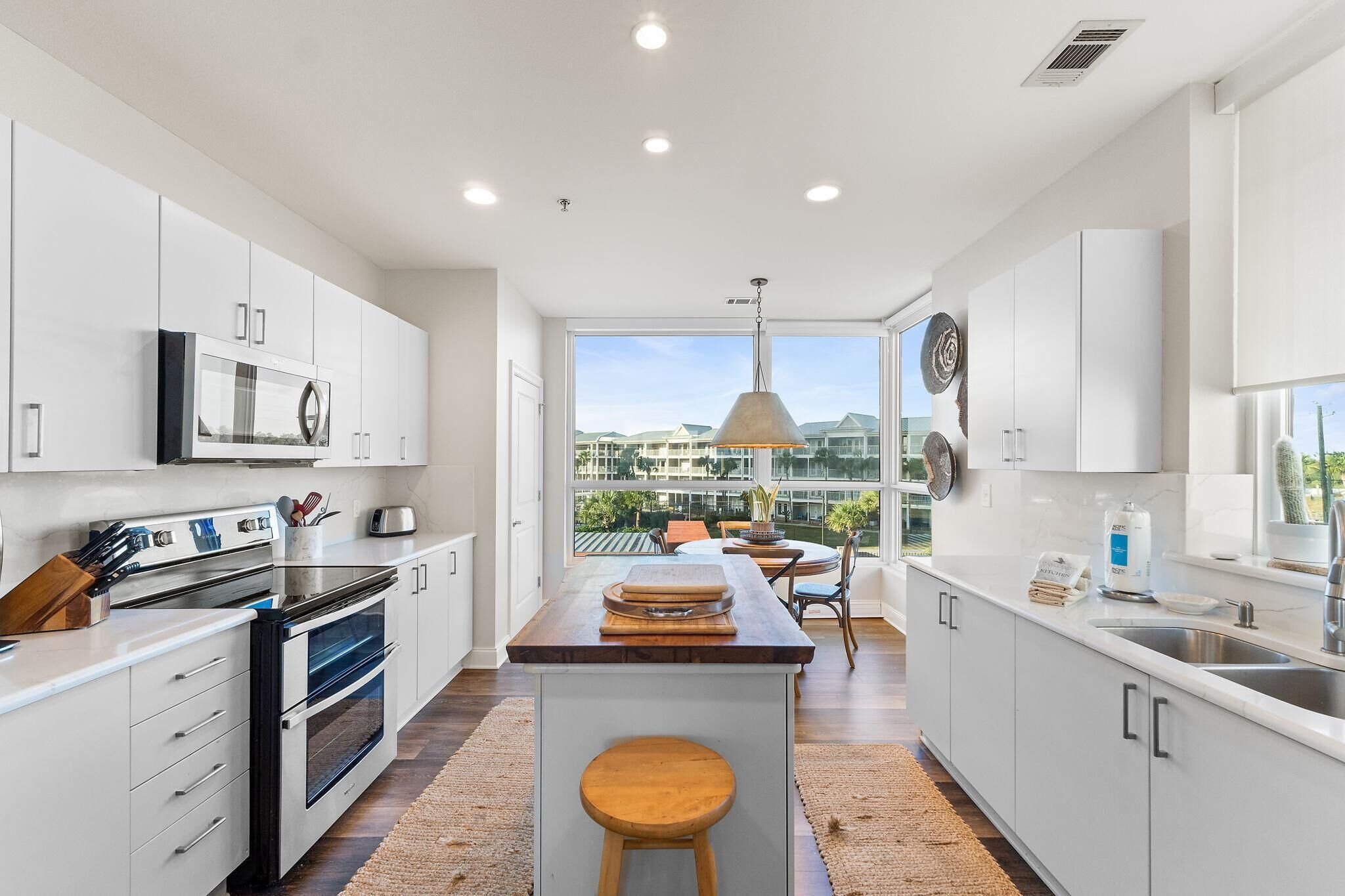 A modern kitchen with white cabinets, a central island with stools, a large window with a view of houses, and a pendant light.