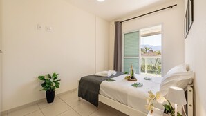 A neatly arranged bedroom with a bed, a potted plant, a bedside table with a lamp, and a window with a view of palm trees. - 200m da praia c piscina e churrasqueira (Bertioga)
