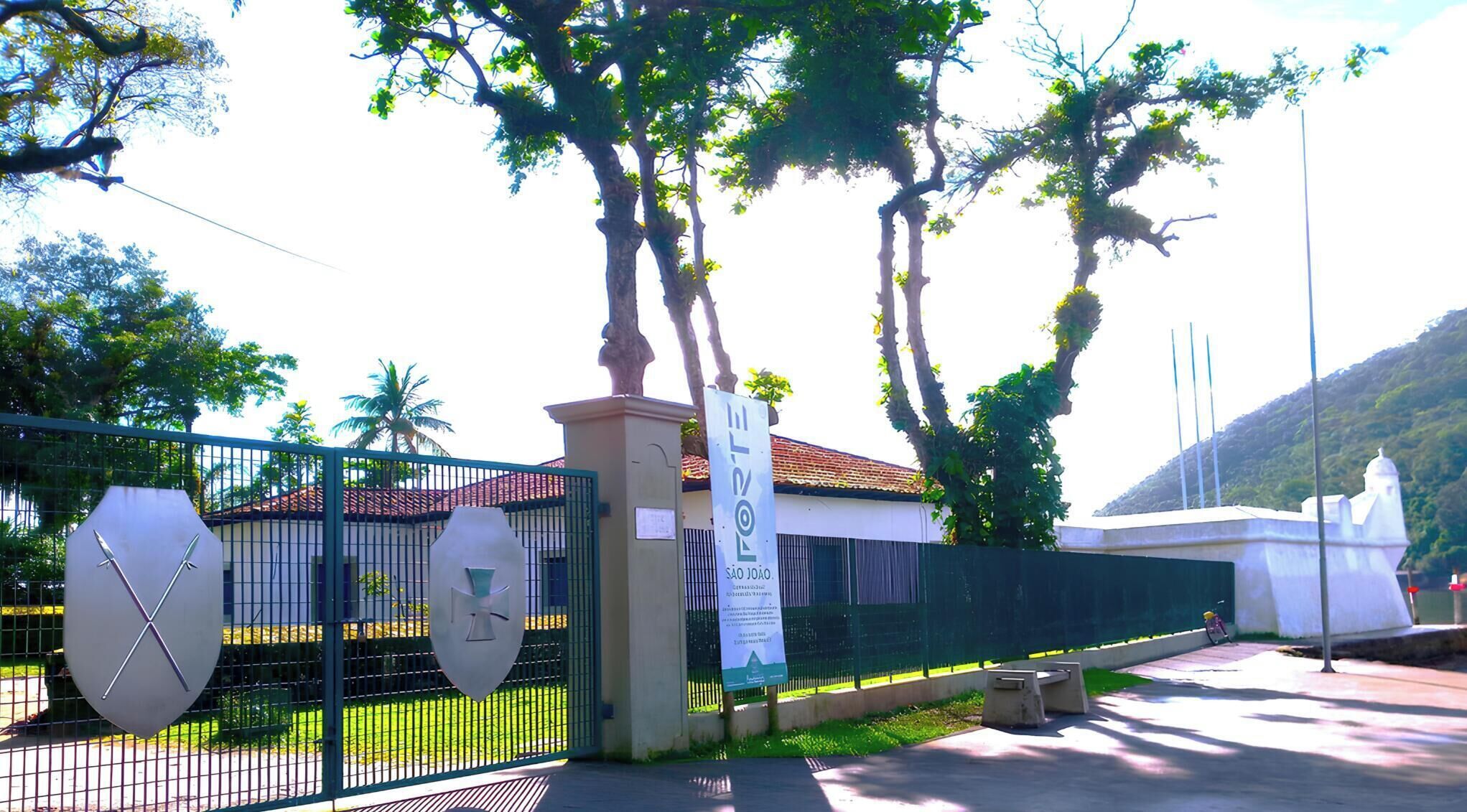 A gated entrance with a sign reading "Forte São João," a white building with a tiled roof, and a statue in the background.