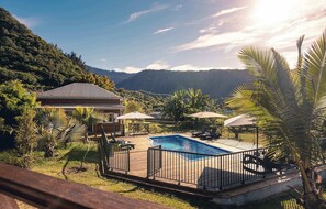 Piscina com deck de madeira, rodeada por palmeiras e um cenário montanhoso.