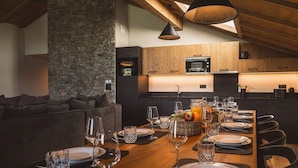 A modern kitchen with wooden cabinets, a stone accent wall, and a dining area set for six with a long wooden table. - Alpine Family Escape with Spa Access (MEGEVE)
