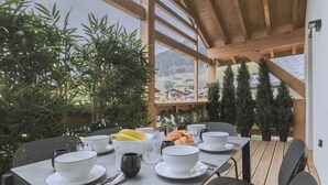 Une table à manger dressée avec des bols, des assiettes, des couverts et des plats, entourée de plantes en pot et offrant une vue sur un paysage montagneux.