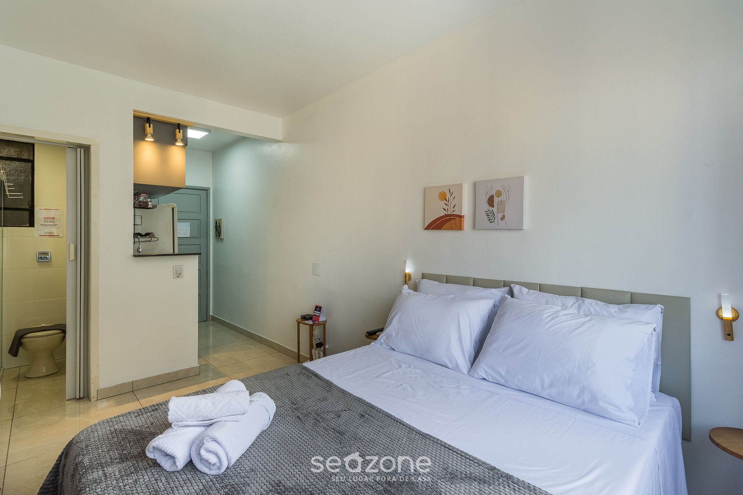 A hotel room with a bed, a bedside table, a nightstand, a wall-mounted light, and a door leading to a bathroom.
