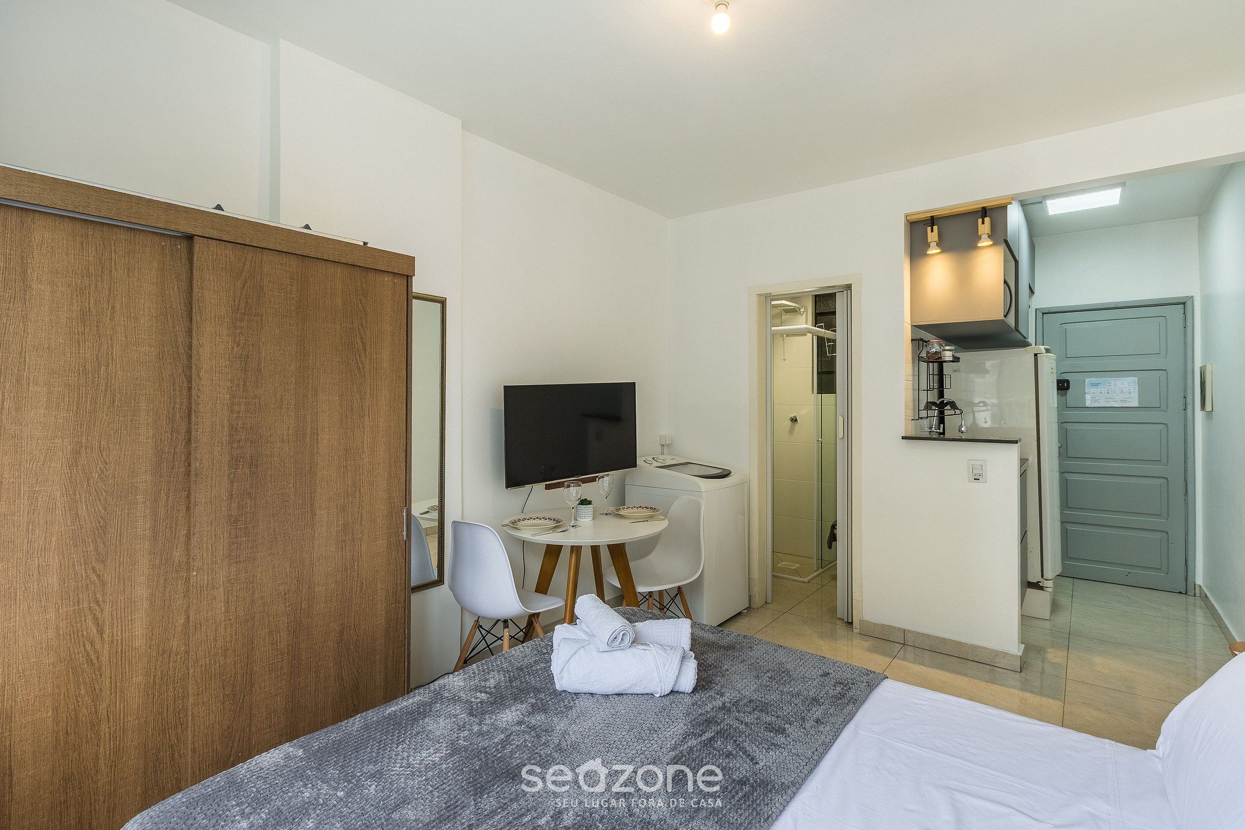 A hotel room with a bed, a TV, a small dining table with chairs, a kitchenette, and a wardrobe.