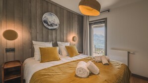 A bedroom with a wooden wall, a bed with white and mustard pillows, a round wall clock, and a view of mountains through a window. - Family Retreat in Peisey-Vallandry (Plan Perisey)