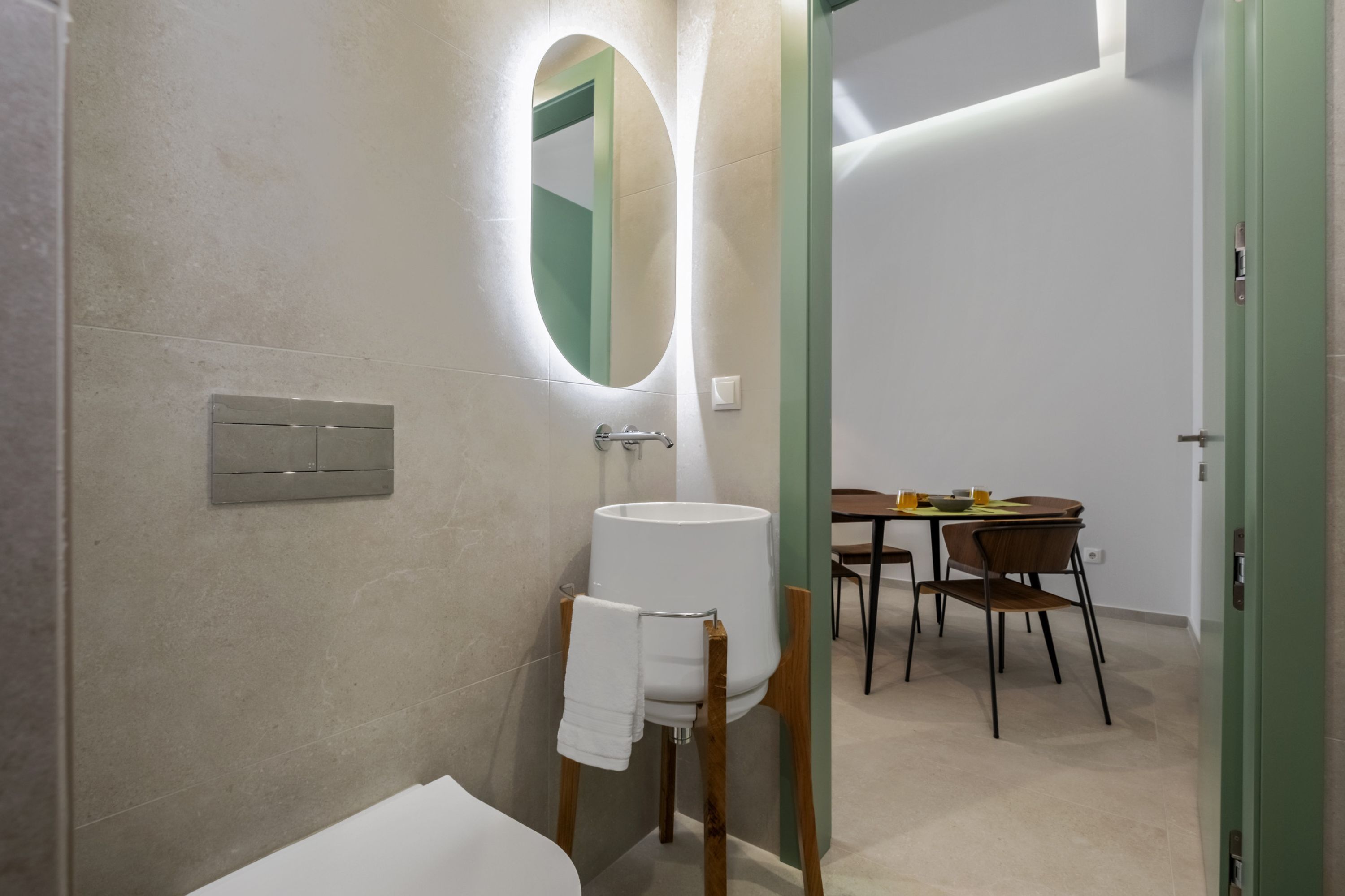 A modern bathroom with a toilet, sink, and mirror. There is a dining area with a table and chairs visible through an open door.