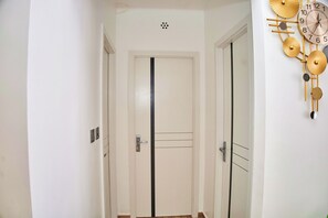 Um corredor com uma porta branca, um relógio fixado na parede e um respiradouro no teto.
