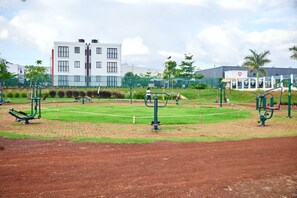 Uma área de ginástica ao ar livre com equipamentos de exercício, um campo de esportes cercado e um edifício ao fundo.