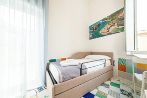 Ein ordentlich bezogenes Bett mit grauer Decke und weißen Kissen, ein farbenfroher Fliesenboden und ein Gemälde einer Küstenlandschaft an der Wand.
