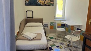 Um quarto com uma cama, uma janela, uma cadeira, uma mesa e um quadro com uma paisagem costeira.