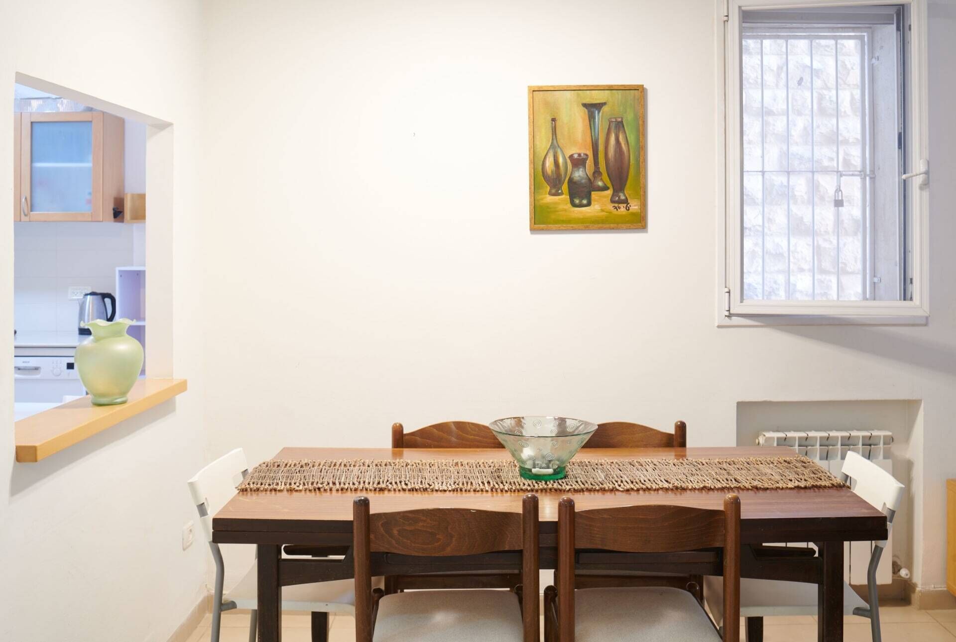 Un comedor con una mesa de madera, dos sillas, un cuenco de vidrio y un cuadro de jarrones en la pared.