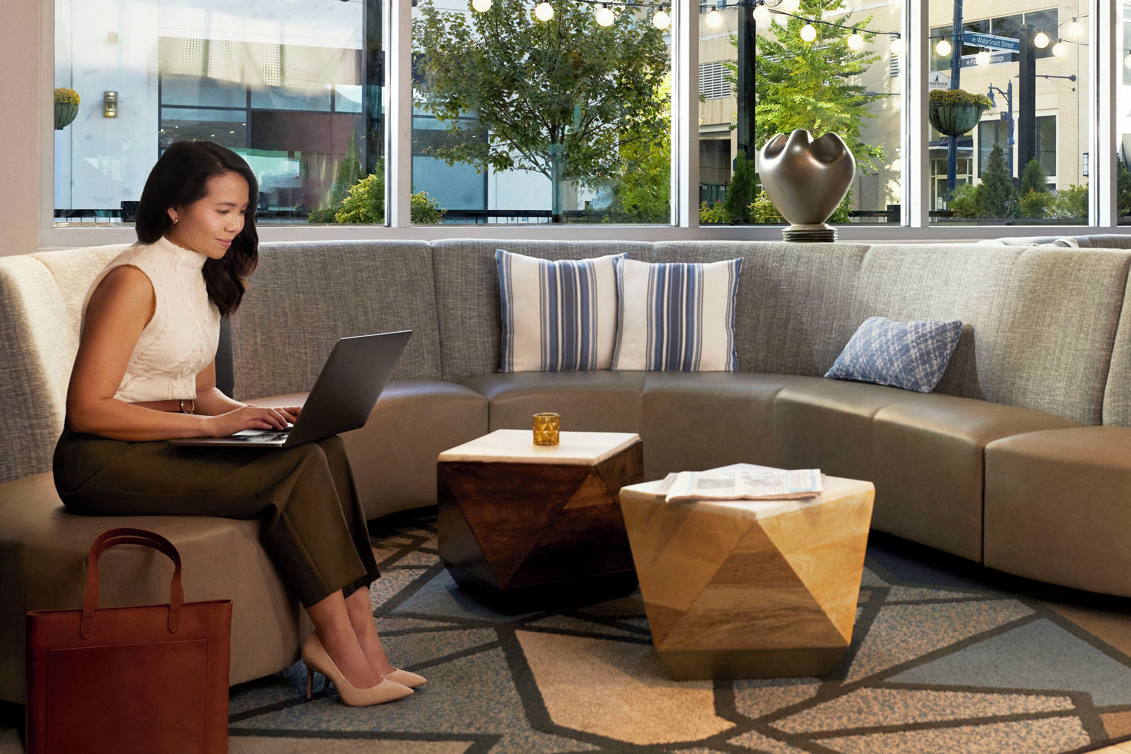 A woman using a laptop on a sofa in a modern room with a geometric patterned carpet, a small wooden side table, and a view of a building and trees outside.