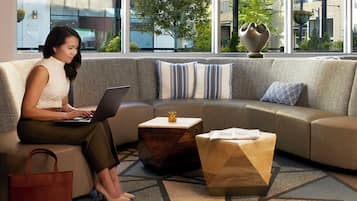A woman using a laptop on a sofa in a modern room with a geometric patterned carpet, a small wooden side table, and a view of a building and trees outside.