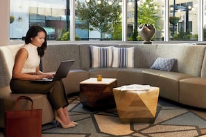 A woman using a laptop on a sofa in a modern room with a geometric patterned carpet, a small wooden side table, and a view of a building and trees outside. - Hyatt Place Seoul Pangyo (Seongnam)