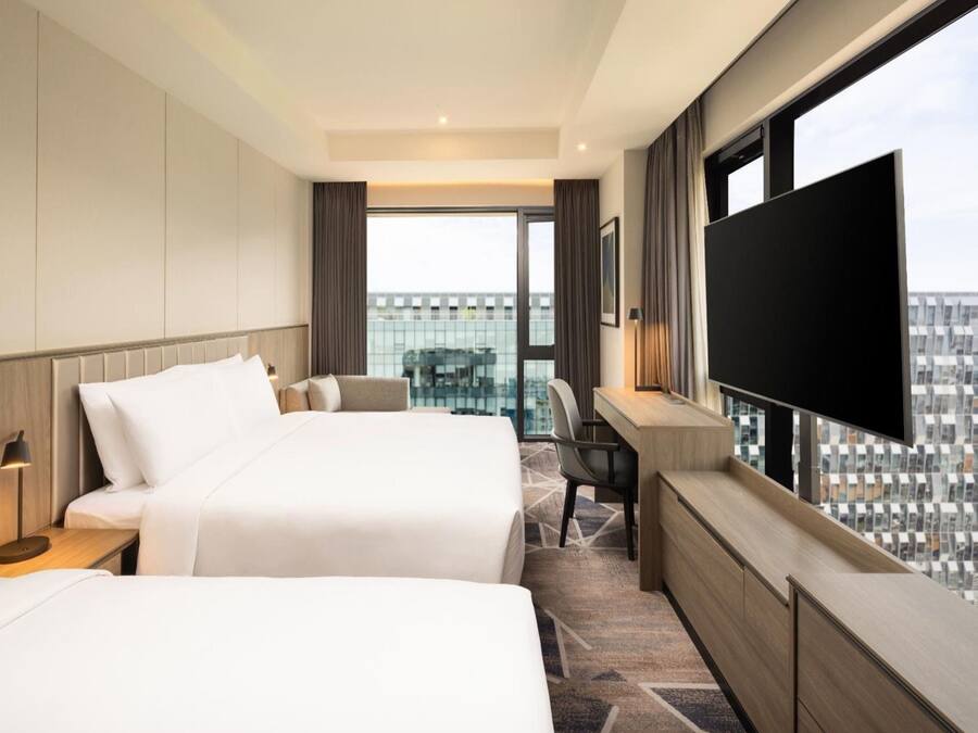 A hotel room with two beds, a desk, a chair, a TV, and a large window with a city view.