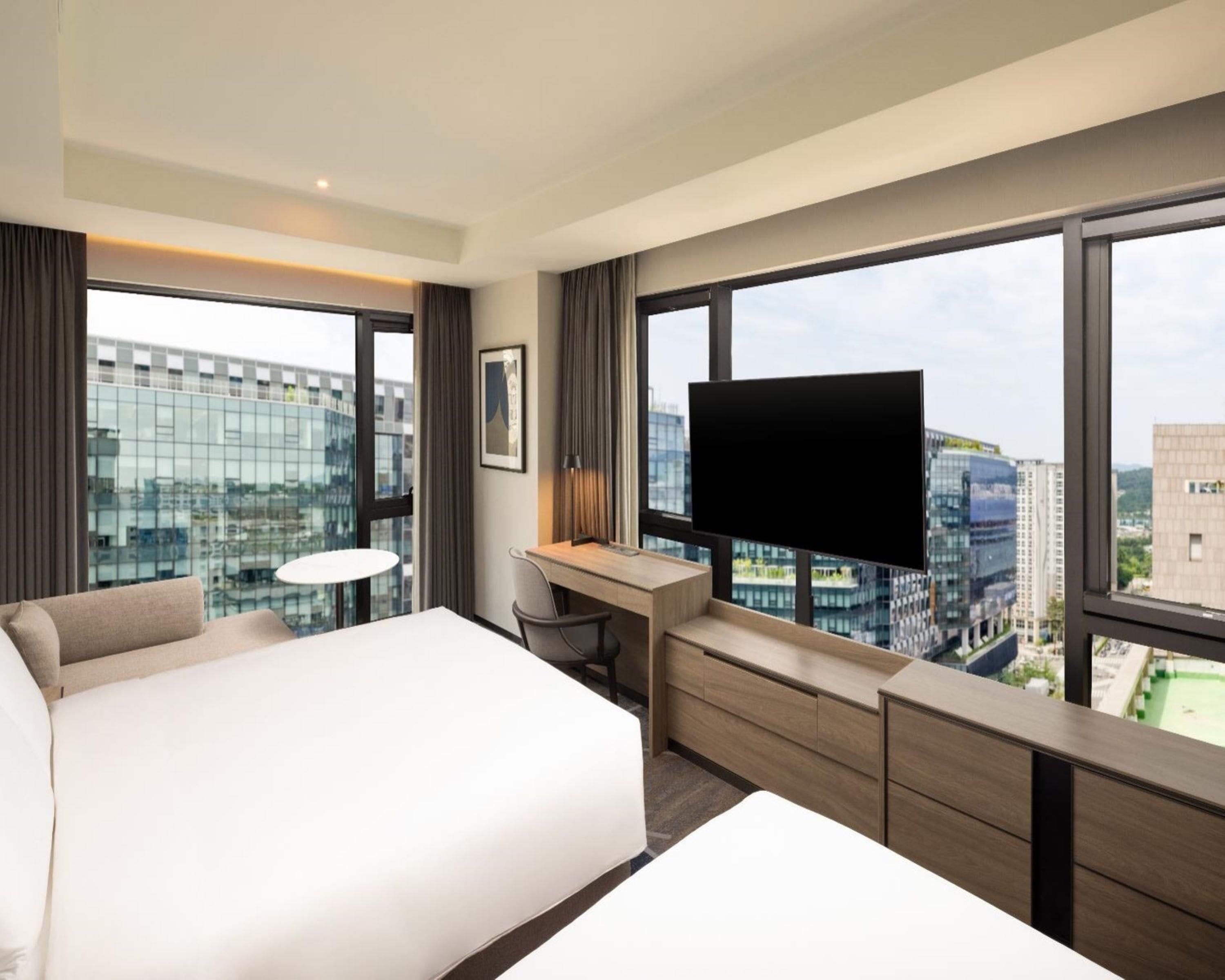 Quarto de hotel com uma cama grande, escrivaninha, cadeira, televisão e vista para a cidade através de amplas janelas.