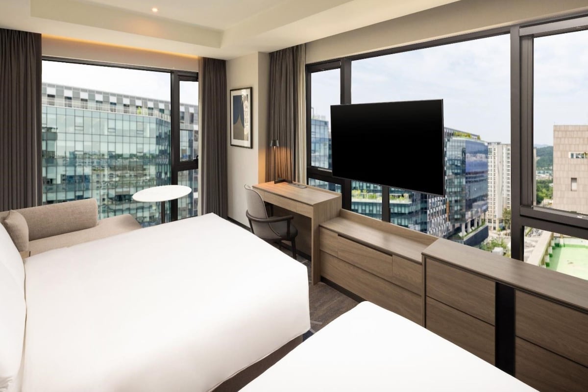 Habitación de hotel con una cama grande, un escritorio, una silla, un televisor y vistas a la ciudad a través de amplias ventanas.