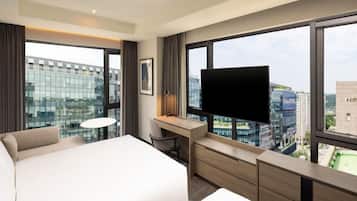 Quarto de hotel com uma cama grande, escrivaninha, cadeira, televisão e vista para a cidade através de amplas janelas.