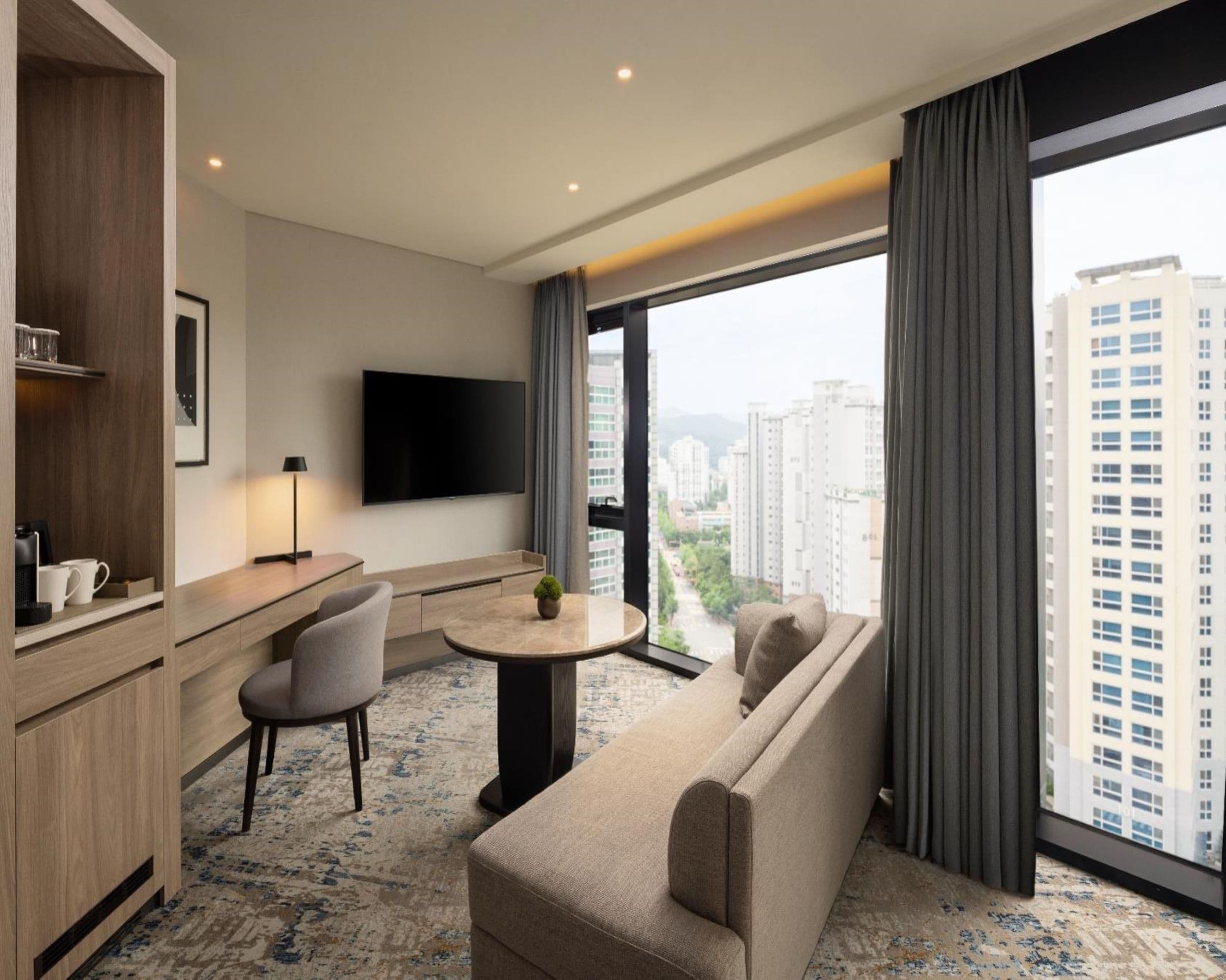 Quarto de hotel moderno com sofá, mesinha redonda, TV de tela plana e uma grande janela com vista para a cidade.