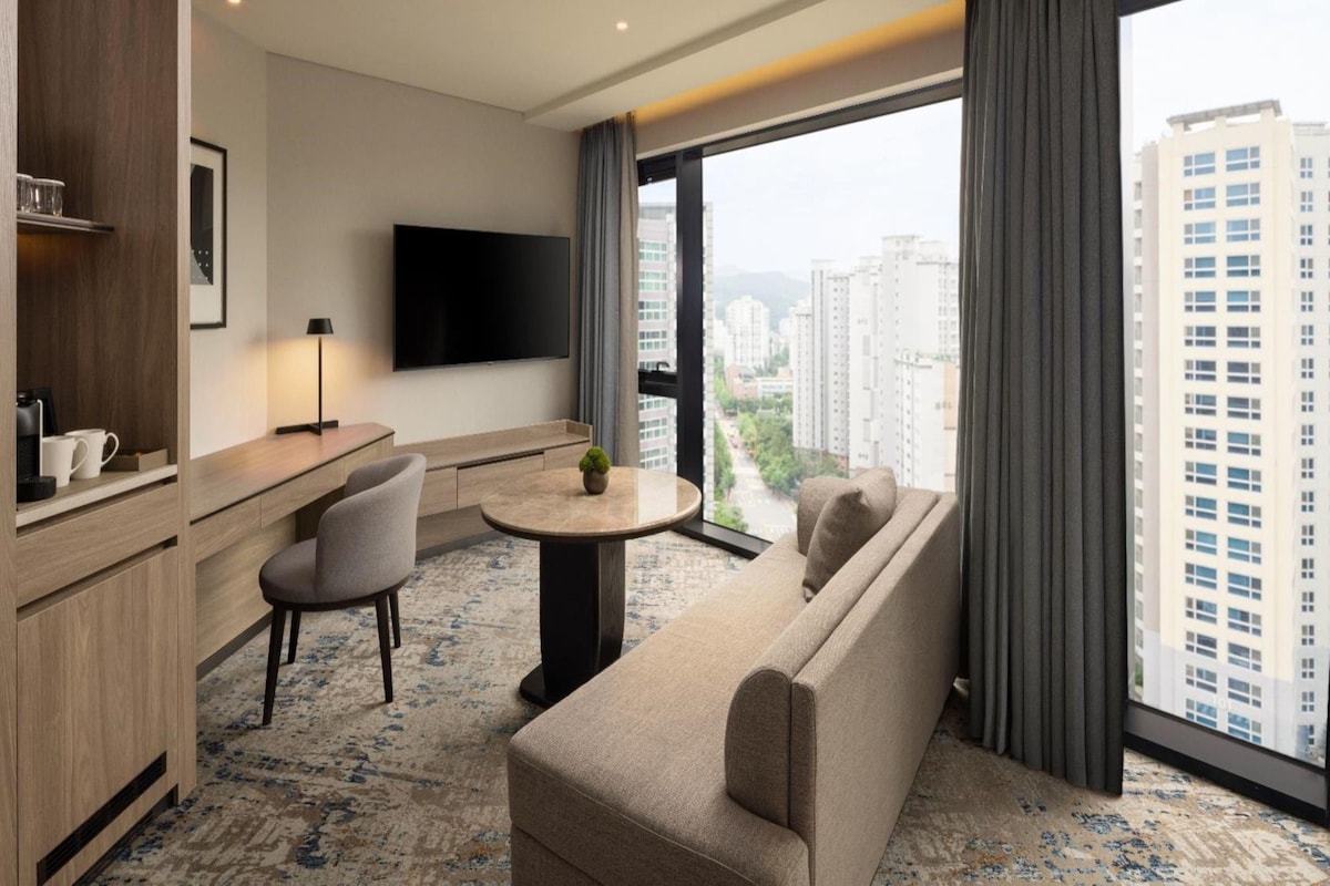 Habitación de hotel moderna con sofá, una pequeña mesa redonda, televisor de pantalla plana y un amplio ventanal con vistas a la ciudad.