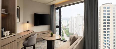Ein modernes Hotelzimmer mit einer Couch, einem kleinen runden Tisch, einem Flachbildfernseher und einem großen Fenster mit Blick auf die Stadt.