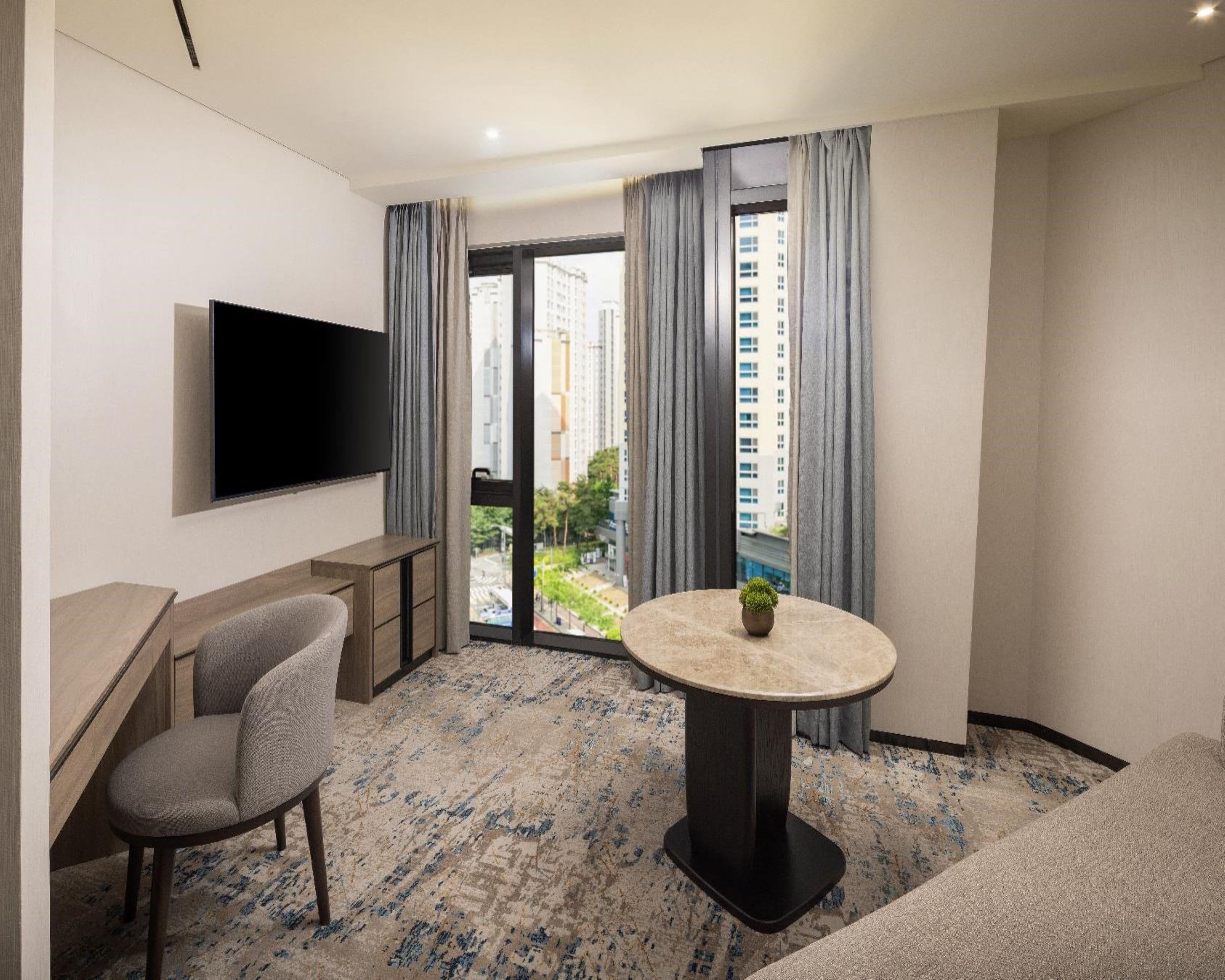 A hotel room with a flat-screen TV mounted on the wall, a round table with a potted plant, a sofa, and a view of a cityscape through the window.