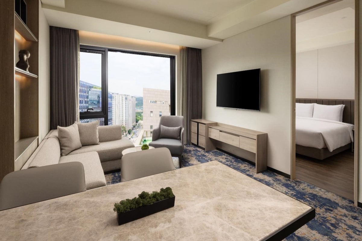 Una habitación de hotel moderna con un conjunto de sofás, una mesa de comedor, un televisor montado en la pared y una cama.