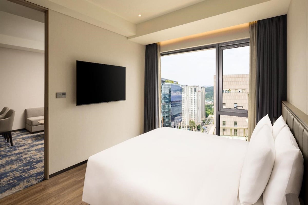 Habitación de hotel con una cama grande, un televisor de pantalla plana montado en la pared, una zona de estar con sofá y sillones, y vistas a la ciudad a través de la ventana.
