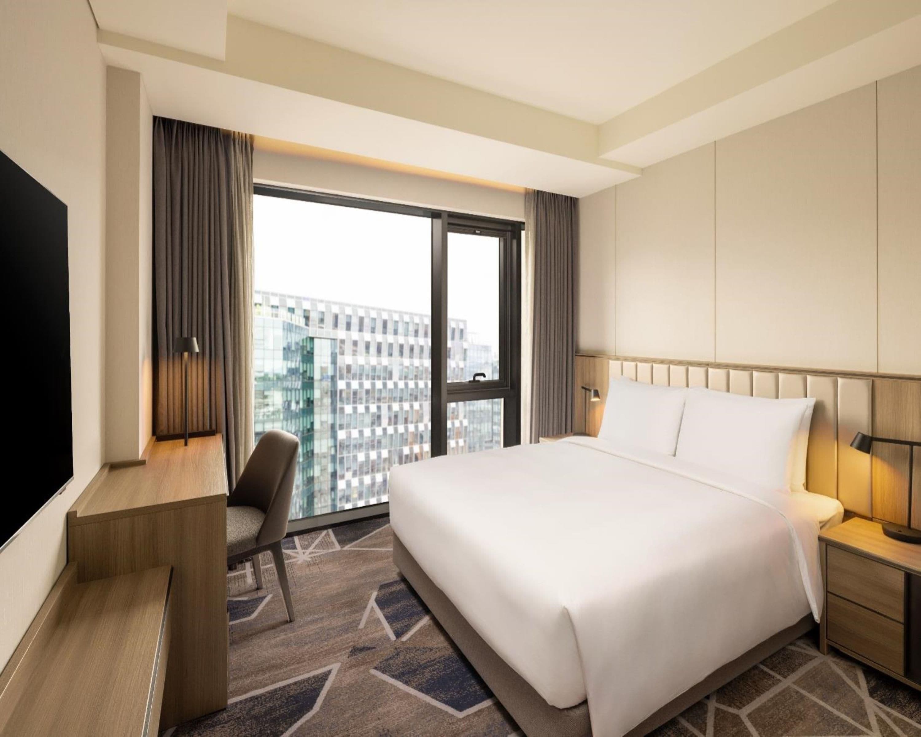 A hotel room with a large bed, a desk, a chair, a TV, and a window with a city view.