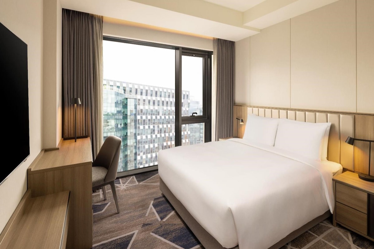 Habitación de hotel con una cama grande, un escritorio, una silla, un televisor y una ventana con vista a la ciudad.